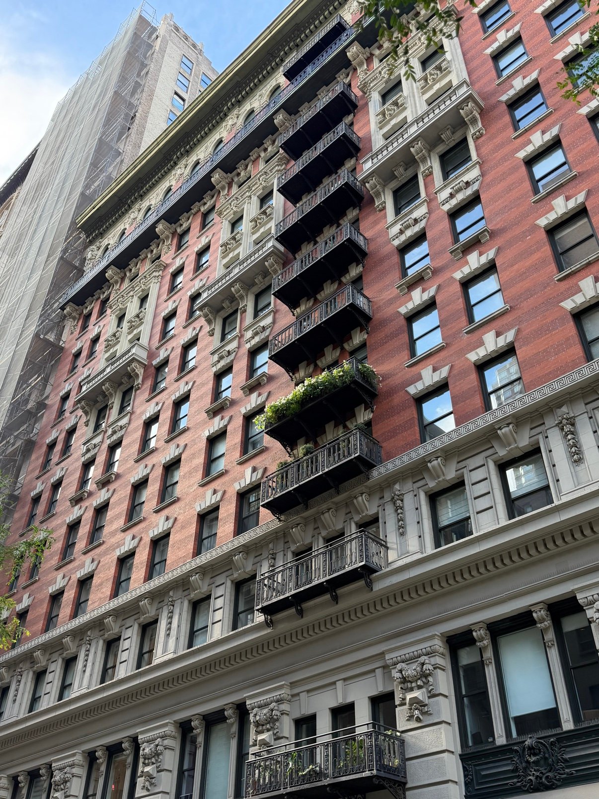 Brick Manhattan apartment building with rows of black iron balconies, one covered in plants